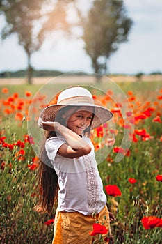 Little longhaired girl in hat posing at field of poppies with  on summer sun. Vertical