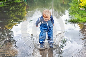 Little cute playful caucasian blond toddler boy enjoy have fun playing jumping in dirty puddle wearing blue waterproof