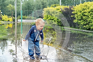 Little cute playful caucasian blond toddler boy enjoy have fun playing jumping in dirty puddle wearing blue waterproof