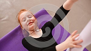 Little cute red-haired girl ballerina performs stretching exercises in ballet school on the background of a group of