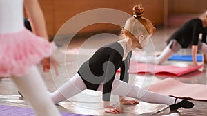 Little cute red-haired girl ballerina performs stretching exercises in ballet school on the background of a group of