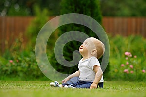 Little cute smiling baby in jeans and white shirt sitting on grass in garden and looking up. Bright green background