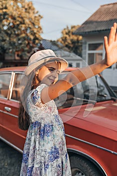 Little cute smiling girl in dress with flowers and white hat standing near red retro car and exetended her hand to cover