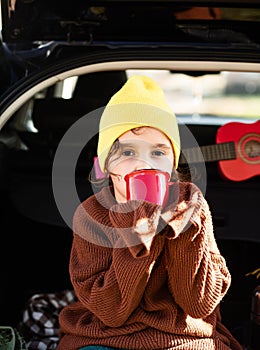 Little cute smiling girl sitting in open car trunk and drinking cocoa. Kid resting with her family in the nature. Autumn season