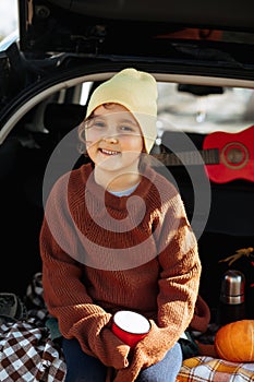 Little cute smiling girl sitting in open car trunk and drinking cocoa. Kid resting with her family in the nature. Autumn season