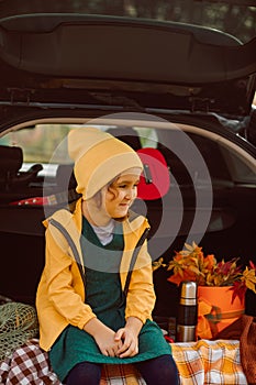 Little cute smiling girl sitting in open car trunk. Kid resting with her family in the nature. Autumn season