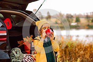 Little cute smiling girl standing near by on open car trunk and drinking cocoa. Kid resting with her family in the nature. Autumn