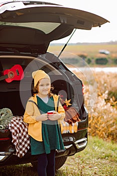 Little cute smiling girl standing near by on open car trunk and drinking cocoa. Kid resting with her family in the nature. Autumn