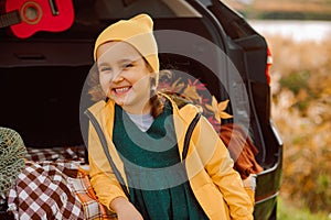 Little cute smiling girl standing near by on open car trunk. Kid resting with her family in the nature. Autumn season