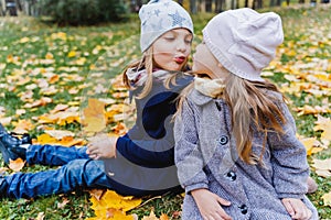 Little cute stylish long hair girls, kissing sisters in autumn coats sitting in multi-colored yellow red maple leaves, foliage.