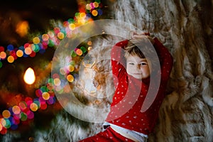 Little cute toddler girl lies under Christmas tree and dreaming of Santa at home, indoors. Traditional Christian