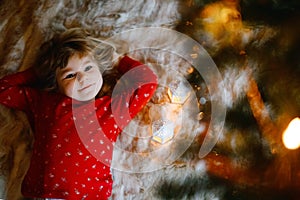 Little cute toddler girl in pajamas under Christmas tree and dreaming of Santa at home, indoors. Traditional Christian