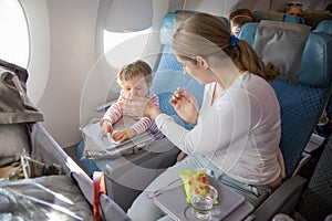 Little cute toddler girl sitting in an airplane in a chair by the porthole, holds a wet towel in her hands and sneezes