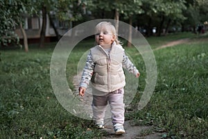 A little cute two-year-old girl in a beige vest and a light suit walks alone in the park. Toddler proudly walks along the path,