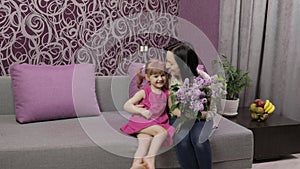 Little daughter child congratulates her mother with bouquet of lilac flowers