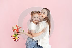 A little daughter gives her mother flowers and a gift for mother& x27;s day on a pink background in the studio. Happy mom