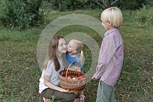 Little daughter kisses her Young mother sitting in clearing near tree. Mom with children and apples for wicker basket