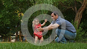 Little daughter takes first steps to meet laughing dad on lawn grass in park. Dad kisses baby on cheek and smill. Slow