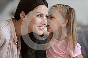 Little daughter telling secret to smiling mother, whispering in ear