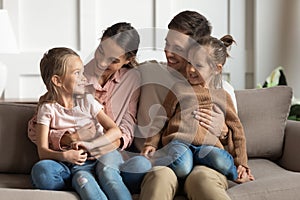 Little daughters sit on parents laps talking resting on couch