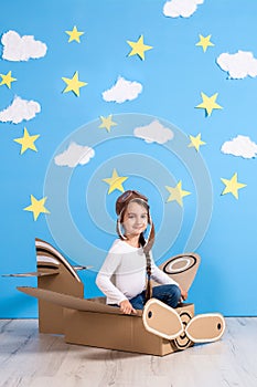 Little dreamer girl playing with a cardboard airplane at the studio with blue sky and white clouds background.