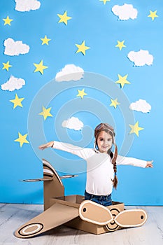 Little dreamer girl playing with a cardboard airplane at the studio with blue sky and white clouds background.