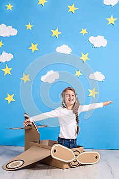 Little dreamer girl playing with a cardboard airplane at the studio with blue sky and white clouds background.