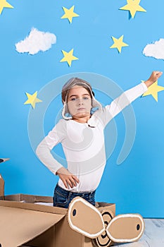 Little dreamer girl playing with a cardboard airplane at the studio with blue sky and white clouds background.