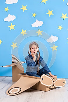 Little dreamer girl playing with a cardboard airplane at the studio with blue sky and white clouds background.