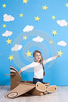 Little dreamer girl playing with a cardboard airplane at the studio with blue sky and white clouds background.