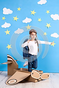 Little dreamer girl playing with a cardboard airplane at the studio with blue sky and white clouds background.