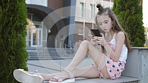 A little elementary school girl sits on a parapet in the background of the school`s campus building and uses a