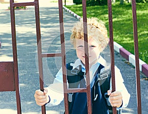 Little elementary school student stands next to his school holding hands for the school fence.