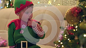 little elf boy put finger in mouth looks pensively at decorated Christmas tree
