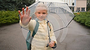 Little European girl wave hand at camera walk skipping umbrella city outside park schoolgirl jumping learner backpack