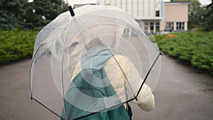 Little European girl wave hand at camera walk skipping umbrella city outside park schoolgirl jumping learner backpack