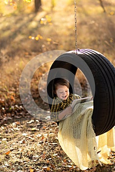 Little fair-haired boy with a smile reads a book in a swing wheel outdoors,