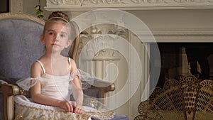 The little fair-haired girl sits on a chair at a fireplace in a white tutu. Darlings small in white packs.