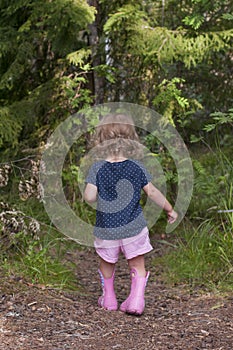 Little fair-haired girl walking along the forest path after the rain. Child in a dark t-shirt, pink shorts and pink rubber boots.