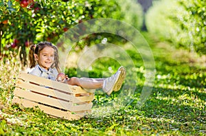 Little farmer girl spending sunny day in garden, having fun, sitting in a crate with apple in hands.