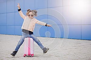 Little fashion curly smiling girl in sunglasses siting on the suitcase and showing peace by fingers. Concept travel