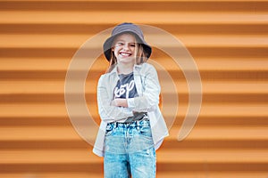 Little fashion-dressed girl portrait sincerely smiling while looking at camera on orange wall background. Urban people living and photo