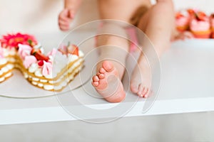 Little feet of baby two years old in pink pants sitting on the white table near the birthday cake and different pink sweets on the