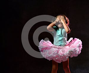 Little female in blue t-shirt and poofy skirt. Laughing, closed her eyes with palms, posing on black background. Close up