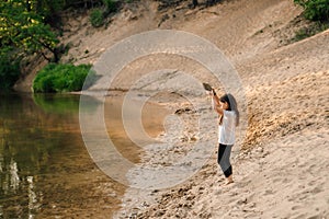 Little female child stand near river on sandy beach in forest and hold clod of grit above head. Girl playing with sand.