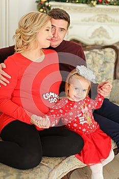 Little female kid sitting with father and pregnant mother near decoreated fireplace.