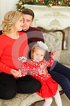 Little female kid wearing red dress sitting with father and pregnant mother near decoreated fireplace.