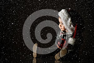 Little female in white hat, gray boots and sparkling suit. Posing sitting on floor in twilight, against black background. Close up