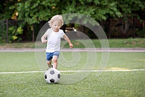 Little football player: blonde child in white shirt and blue shorts running along the green soccer field ready to kick ball.