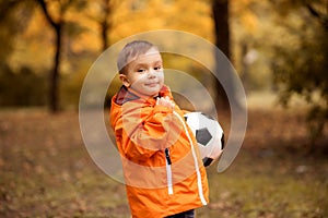Little football player: smiling toddler boy in orange jacket standing half turned and holding soccer ball in autumn park.